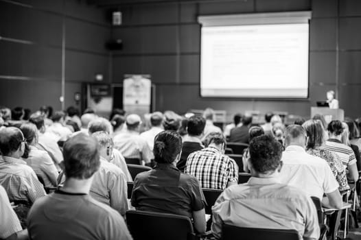 Business and entrepreneurship symposium. Speaker giving a talk at business meeting. Audience in conference hall. Rear view of unrecognized participant in audience.