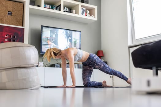 Attractive sporty woman working out at home, doing pilates exercise in front of television in her living room. Social distancing. Stay healthy and stay at home during corona virus pandemic.