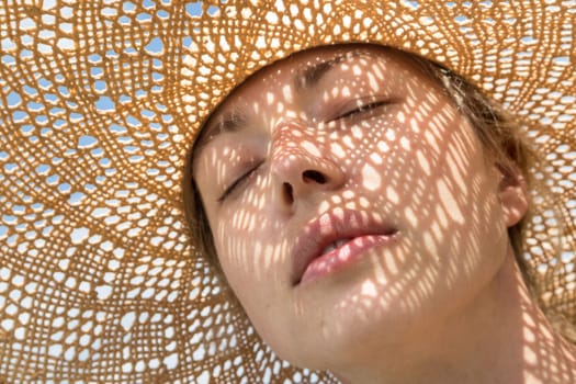 Close-up portrait of woman, closed eyes, wearing straw hat enjoying summer sun. Pattern of shadows falling on her face.