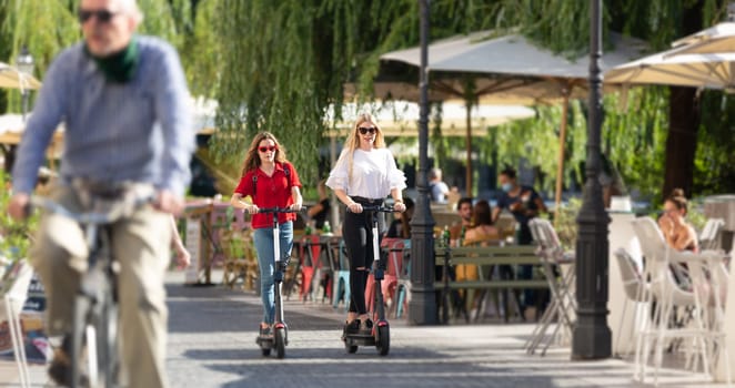 Trendy fashinable teenager girls riding public rental electric scooters in urban city environment. New eco-friendly modern public city transport in Ljubljana, Slovenia.