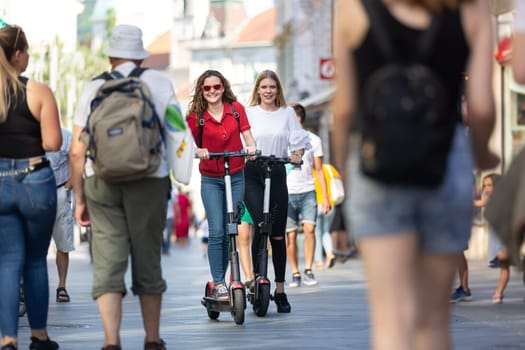 Trendy fashinable teenager girls riding public rental electric scooters in urban city environment. New eco-friendly modern public city transport in Ljubljana, Slovenia.