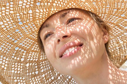 Close-up portrait of woman wearing straw hat enjoying summer sun. Pattern of shadows falling on her face.