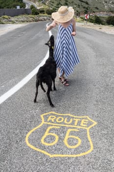 Young attractive female traveler wearing striped summer dress and straw hat standing on an endless straight empty road in the middle of nowhere on Route 66 road and feeding black sheep.