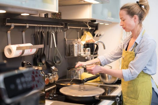 Stay at home housewife woman cooking in kitchen, stir frying dish in a saucepan, preparing food for family dinner. Blonde caucasian model in her 30is, real cooking not looking in camera.