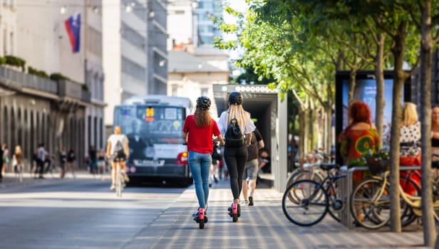 Rear view of trendy fashinable teenager girls riding public rental electric scooters in urban city environment. New eco-friendly modern public city transport in Ljubljana, Slovenia.