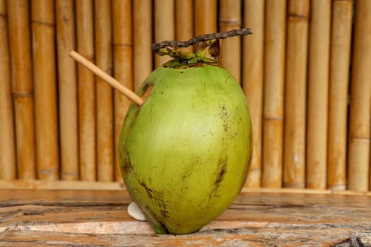 Fresh young coconut with bamboo straw on table with massive wooden board. Healthy natural refreshment full of energy and vitamins. In the background wall of bamboo trunks. Nice tropical concept.