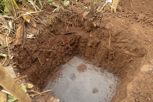 Square, one meter deep hole in the ground with concrete bottom. Hole for a concrete foundation in clay ground. Building foundations for building a reservoir for a family house in a tropical jungle.