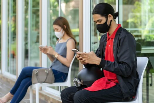 Asian customers wear face mask using mobile phone while sit in waiting social distance queue in front of restaurant with free copyspace for editing. New normal restaurant concept.