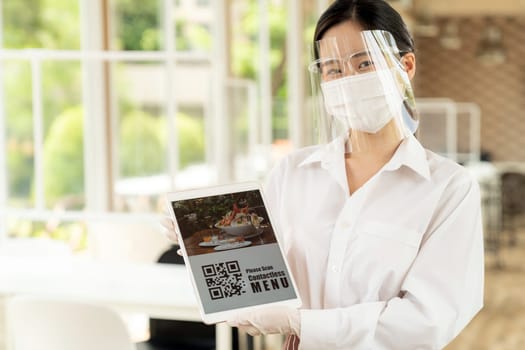 Asian waitress with face mask and face shield hold digital tablet with QR code for customer to scan for online contactless menu. Online contactless and technology concept for new normal restaurant.