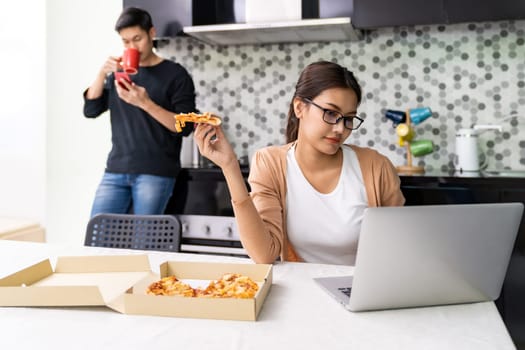 Asian woman work from home in the kitchen and take away pizza while asian man drink coffee in background while city lockdown from coronavirus covid-19 pandemic. New Normal lifestyle working from home.