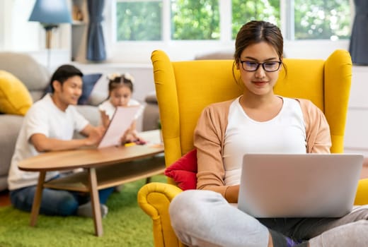 Asian working mom work from home in living room with background of father and daughter reading tale story book while city lockdown. Diversit concept of working mom and family dad and girl.