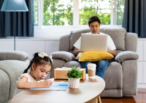 Asian girl drawing in living room with his father working from home with laptop notebook in background while city lockdown from coronavirus covid-19 pandemic. New Normal lifestyle working from home.