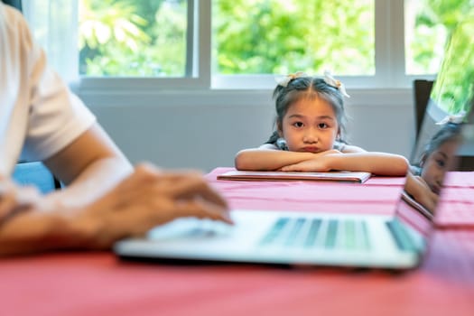 Asian upset girl looking and waiting her father working in dining room. Dad has to working from home while city lockdown from coronavirus covid-19 pandemic. Domestic life dad and daughter concept.