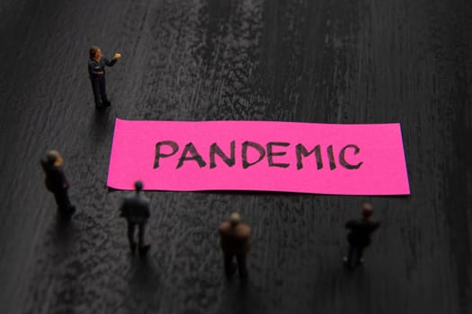 Strategy meeting with miniature figurines posed as business people standing around post-it note with Pandemic message, minimalist abstract concept image with focus on text