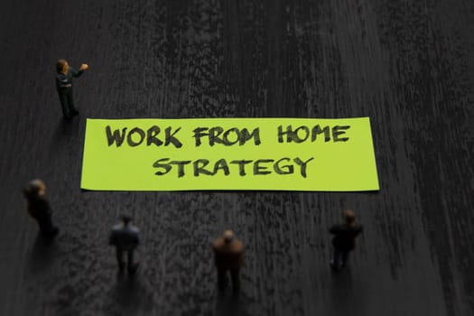 Strategy meeting with miniature figurines posed as business people standing around post-it note with Work From Home Strategy message, minimalist abstract concept image with focus on text