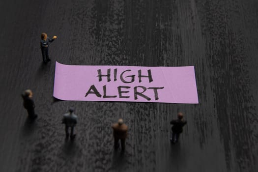 Strategy meeting with miniature figurines posed as business people standing around post-it note with High Alert message, minimalist abstract concept image with focus on text