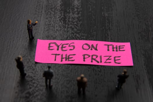 Strategy meeting with miniature figurines posed as business people standing around post-it note with Eyes On The Prize message, minimalist abstract concept image with focus on text