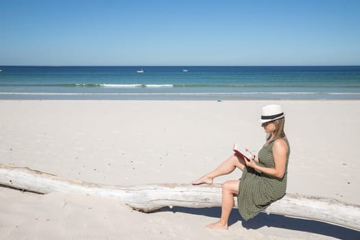 Middle-aged blonde woman sitting on a tree trunk n the beach reading a book with the sea on the background