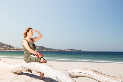 A middle-aged blonde woman sitting on a tree trunk at the beach, protecting herself from the sun with her hand to look in the distance and with a book on her legs