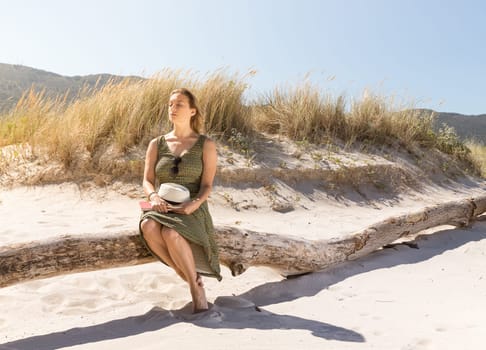 A middle-aged blonde woman sitting thoughtfully on a tree trunk at the beach with a hat and a book on her legs
