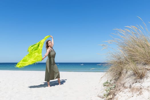 A middle-aged blonde woman holding a green sarong in the wind on the beach with the sea in the background