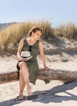 A middle-aged blonde woman sitting thoughtfully on a tree trunk at the beach with a hat and a book on her legs