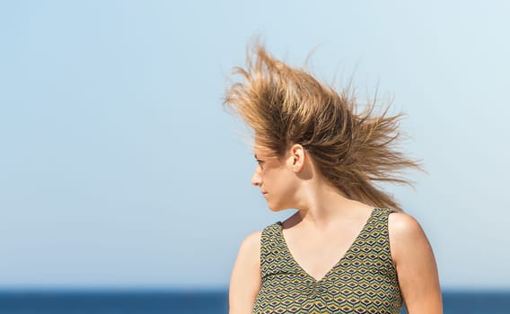 Close-up of a middle-aged blonde woman in a green dress and hair shaking in the wind, with the sea in the background.