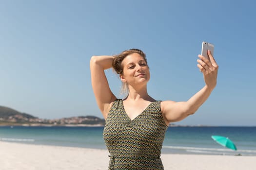 Middle-aged blonde woman in a green dress making a selfie on the beach with the sea in the background
