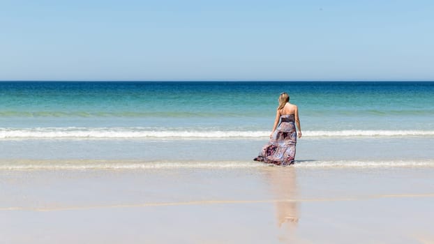 Middle-aged blonde woman in a long dress relaxes on the beach facing the sea