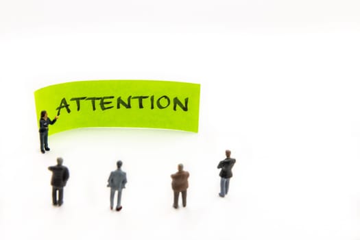 Strategy meeting with miniature figurines posed as business people standing around post-it note with Attention handwritten message on white background, minimalist abstract concept with focus on text