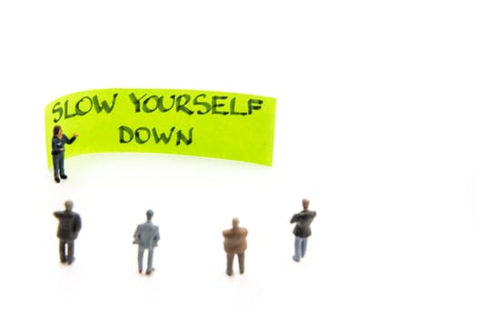 Meeting with miniature figurines posed as business people standing around post-it note with Slow Yourself Down handwritten message on white background, minimalist abstract concept with focus on text