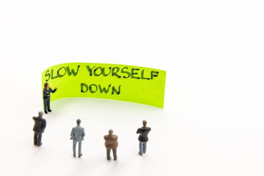 Meeting with miniature figurines posed as business people standing around post-it note with Slow Yourself Down handwritten message on white background, minimalist abstract concept with focus on text