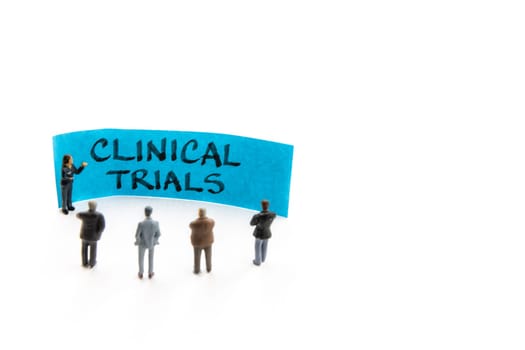 Meeting with miniature figurines posed as business people standing around post-it note with Clinical Trials handwritten message in background, minimalist abstract concept with focus on text