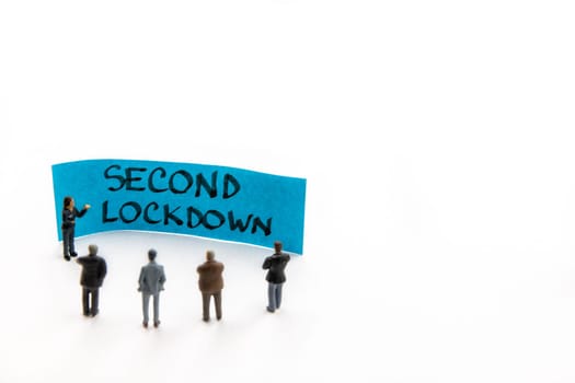 Meeting with miniature figurines posed as business people standing around post-it note with Second Lockdown handwritten message in background, minimalist abstract concept with focus on text
