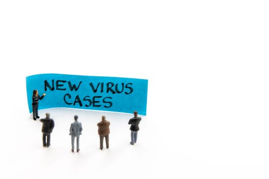 Meeting with miniature figurines posed as business people standing around post-it note with New Virus Cases handwritten message in background, minimalist abstract concept with focus on text
