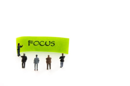 Meeting with miniature figurines posed as business people standing around post-it note with Focus handwritten message in background, minimalist abstract concept with focus on text