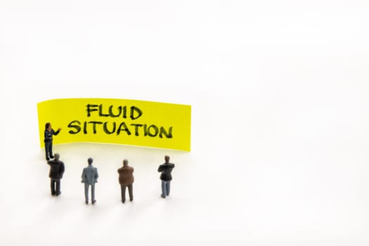Meeting with miniature figurines posed as business people standing around post-it note with Fluid Situation handwritten message in background, minimalist abstract concept with focus on text
