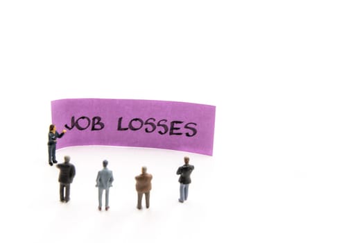 Meeting with miniature figurines posed as business people standing around post-it note with Job Losses handwritten message in background, minimalist abstract concept with focus on text