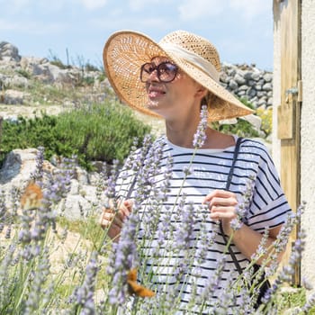 Beautiful blonde young female traveler wearing straw sun hat enjoying summer on Mediterranean cost strolling among lavander flowers on traditional costal village garden.