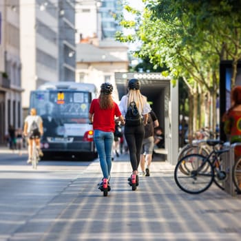 Rear view of trendy fashinable teenager girls riding public rental electric scooters in urban city environment. New eco-friendly modern public city transport in Ljubljana, Slovenia.
