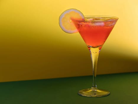Red cocktail in a glass with a lemon slice on a green and yellow background