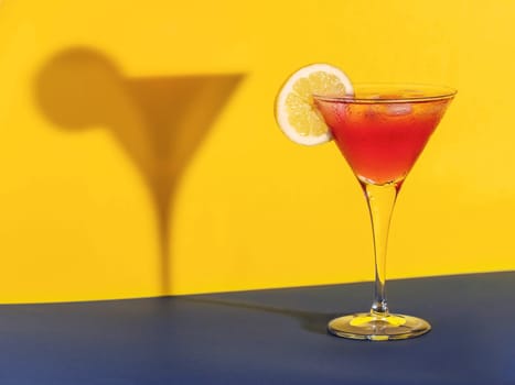 Red cocktail in a glass shaping its shadow with a lemon slice on a blue and yellow background