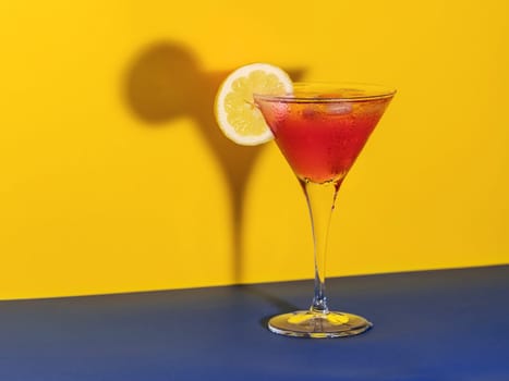 Red cocktail in a glass shaping its shadow with a lemon slice on a blue and yellow background