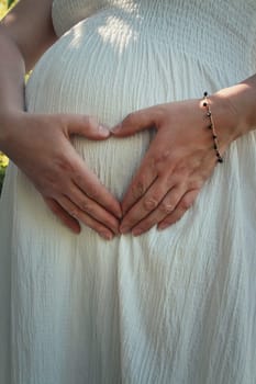 Closeup cute composition with a pregnant young woman wearing white dress makes a heart shaped symbol with her hands on her belly. Happy mother waiting for a baby. Family love concept, motherhood.
