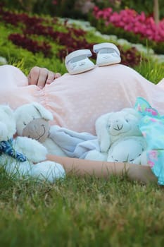 Cute composition with a pregnant young woman laying outdoors on the grass in the park surrounded by multiple baby stuffed toys and newborn shoes on her belly. Happy mother and family waiting for child