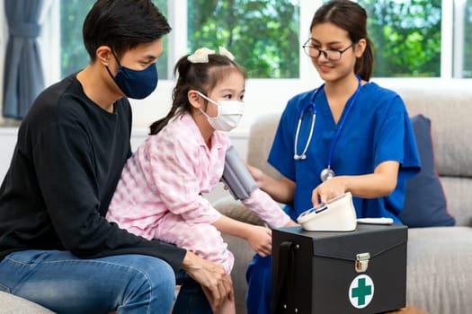 Asian doctor take blood pressure measurement to little girl child sit with her father in living room while doctor visit home. Home healthcare delivery concept.