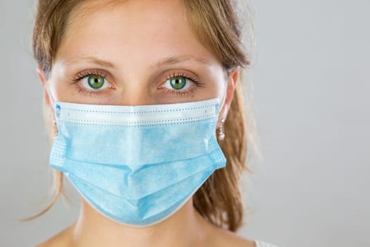 Young woman with green eyes wearing blue surgical face mask and facing camera in studio. Blond lady staring from front with protection over face to prevent infection.