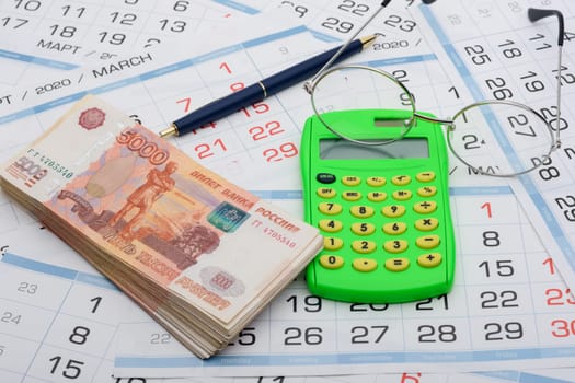 A bundle of money, calculator, glasses and a ballpoint pen lies on the background from the calendar