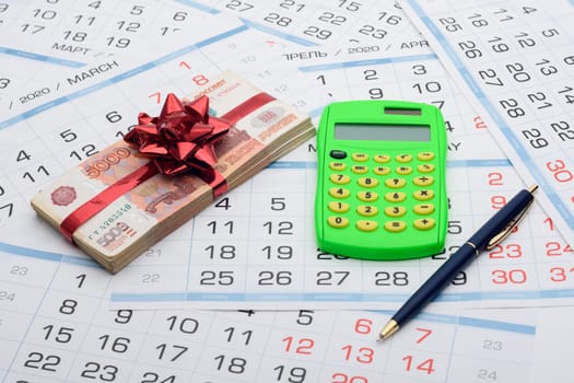 On the background from the calendar lies a bundle of money with a red bow, a calculator and a pen