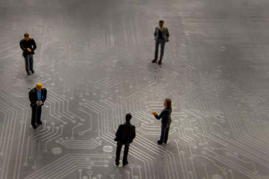 Miniature figurines posed as business people in a meeting over abstract futuristic circuit board, future business concept